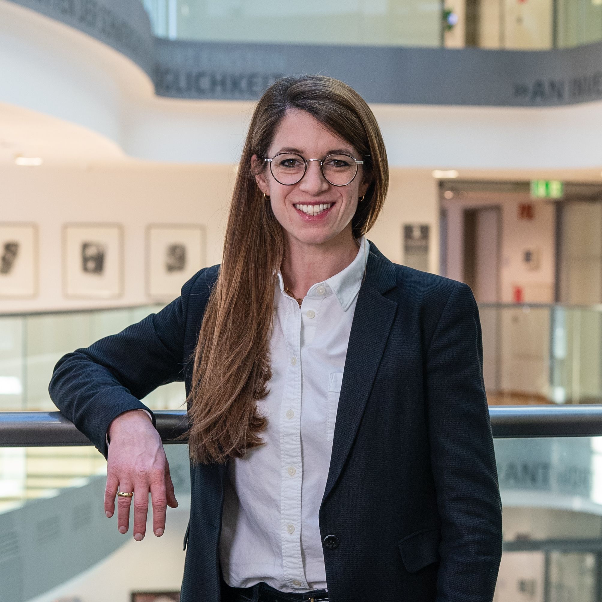A smiling woman with long brown hair, wearing glasses, a white button-down shirt, and a dark blazer, stands leaning on a railing in a modern building atrium with abstract art and text on the walls in the background.